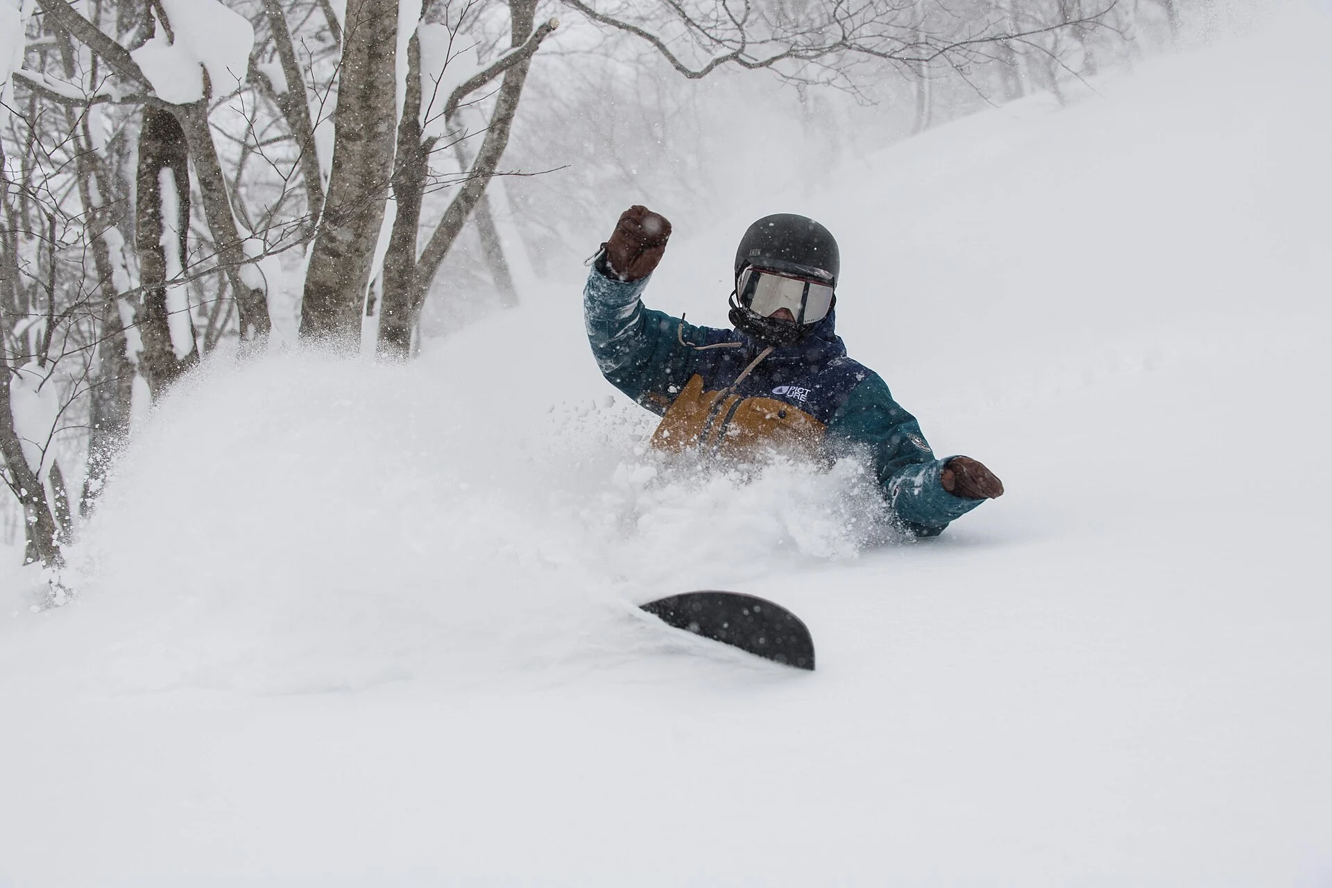 A snowboarder in helmet, goggles, shell jacket and gloves riding deep powder in a Japanese tree run.